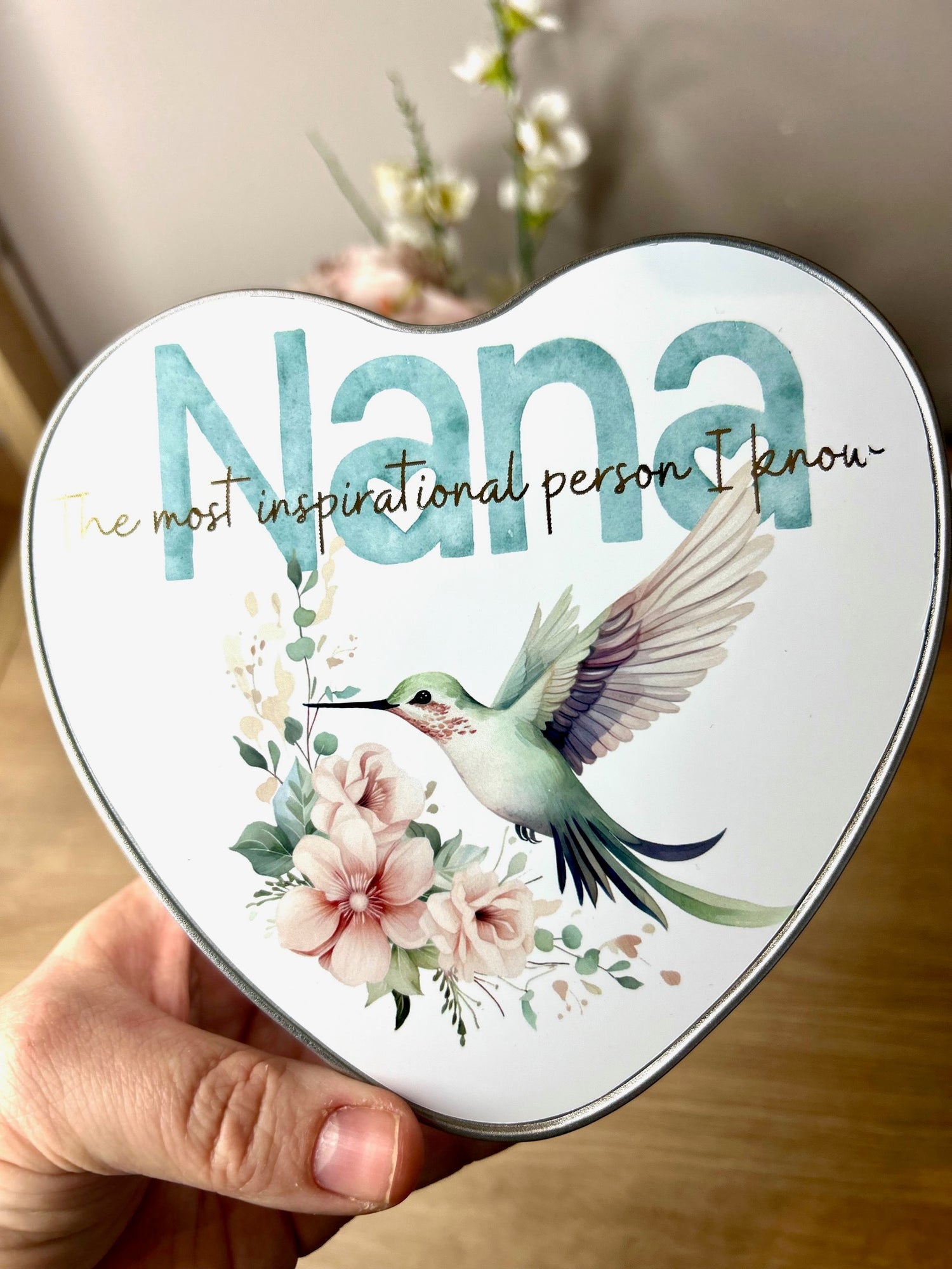 An aluminium heart shaped tin with a picture of a humming bird with flowers on the front and the word Nana