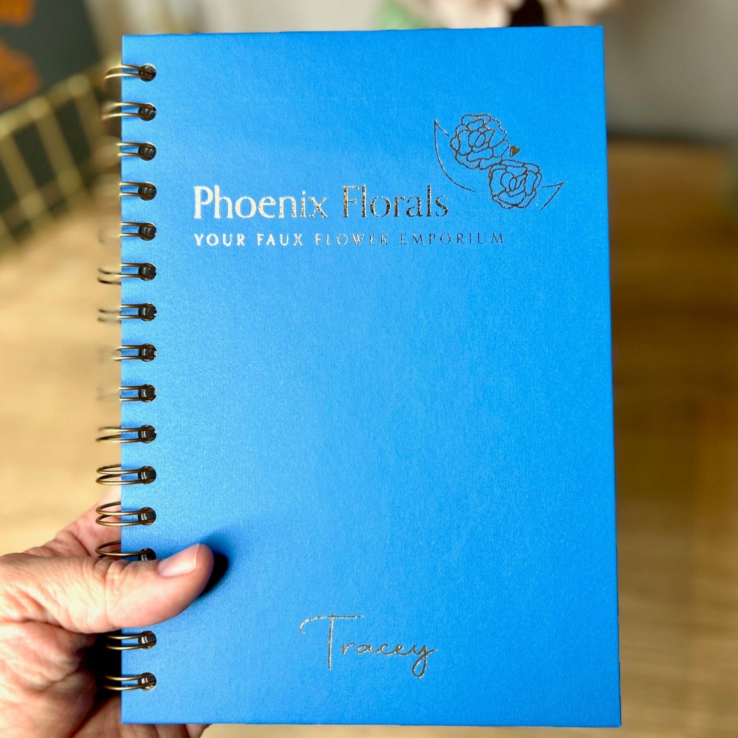 A hardback A5 notebook in Sea Blue that says 'Phoenix Florals your faux flower emporium' with the name 'Tracey' underneath and an image of a flower all in gold ,It is a business logo