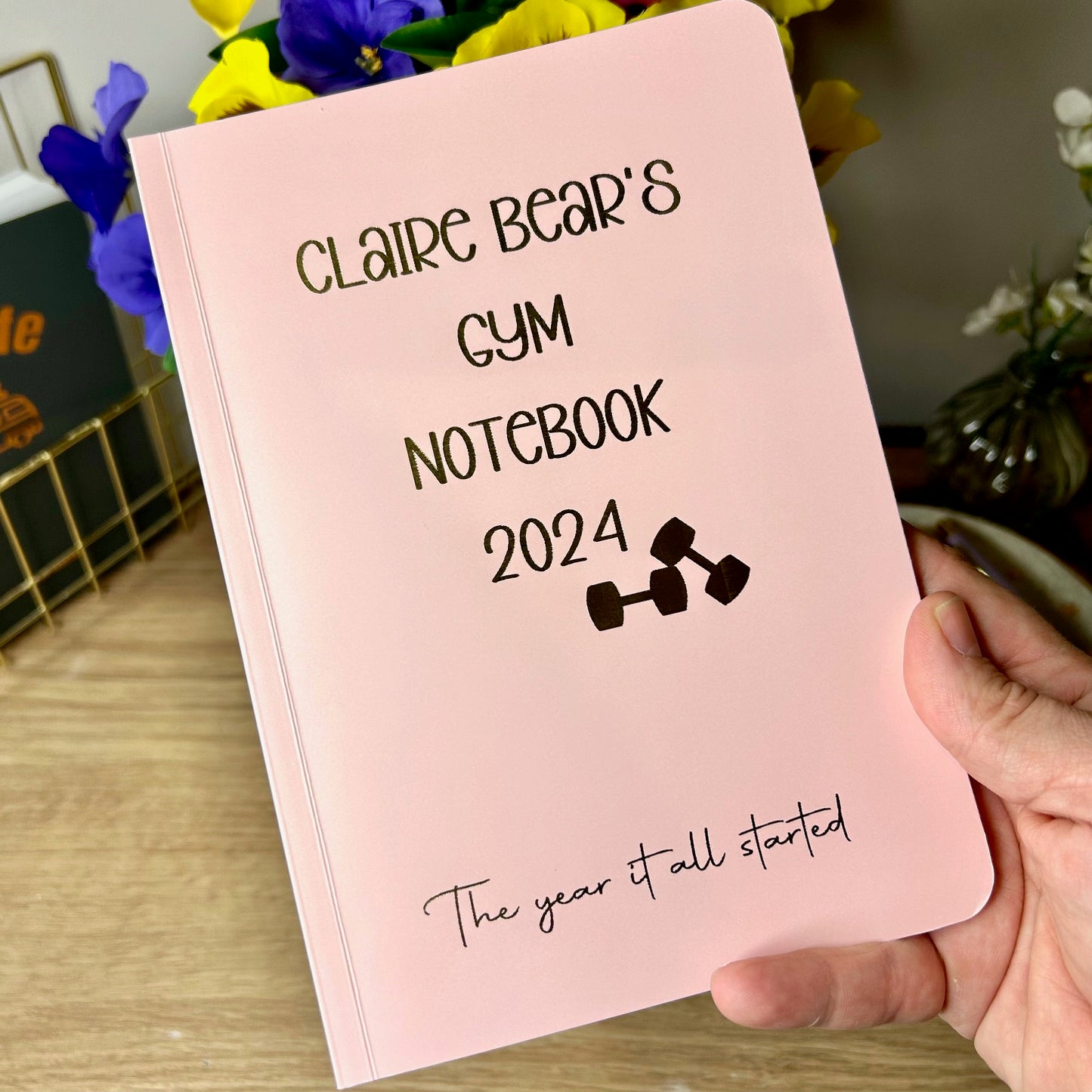 A Poweder pink as softback notebook that says 'Claire bears gym notebook 2024' in gold foil