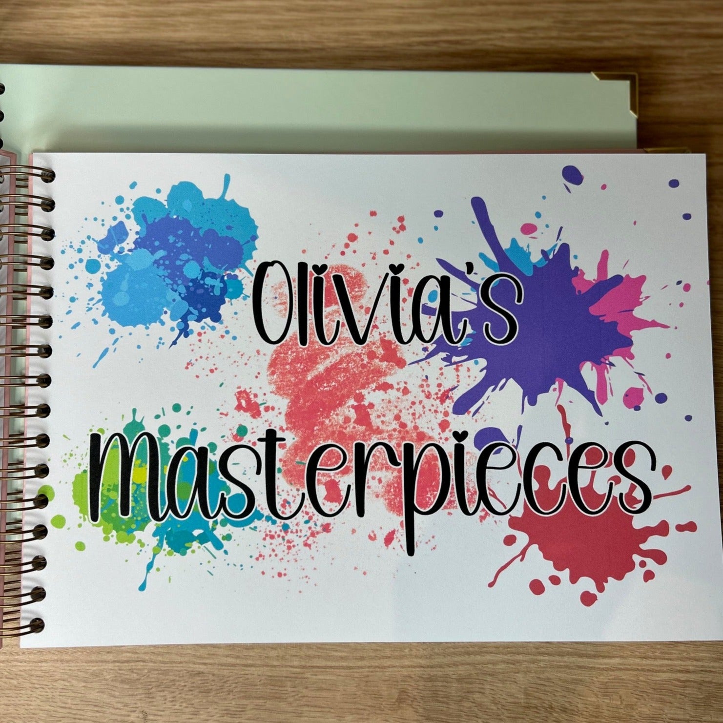 The inside page of the Masterpieces memory book. The page says 'Olivia's Masterpieces in black witing surrounded with multicoloured paint splats