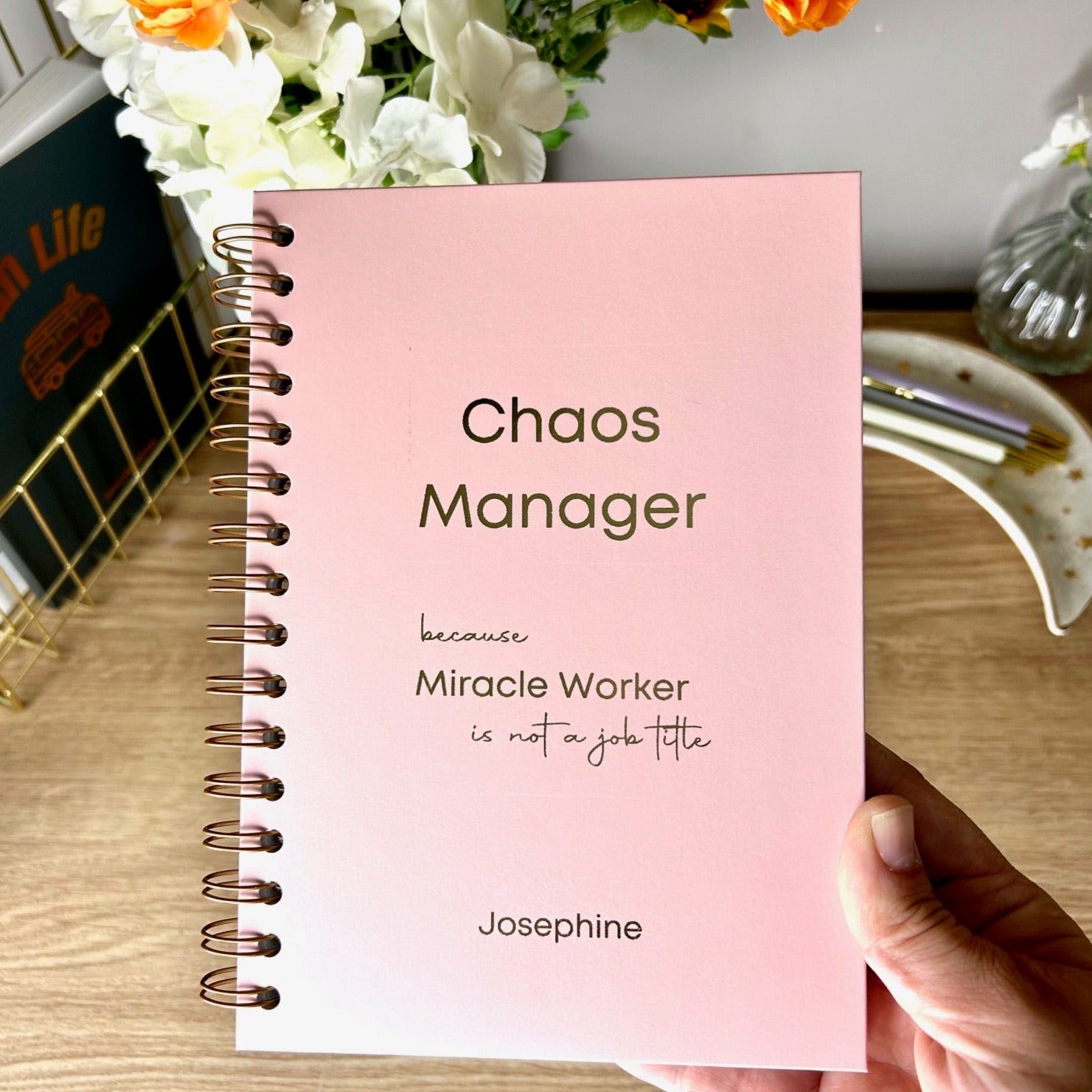 A hardback A5 notebook in Powder Pink that says 'Chaos manager because miracle worker is not a job title' with the name 'Josephine' below, all in gold foil 