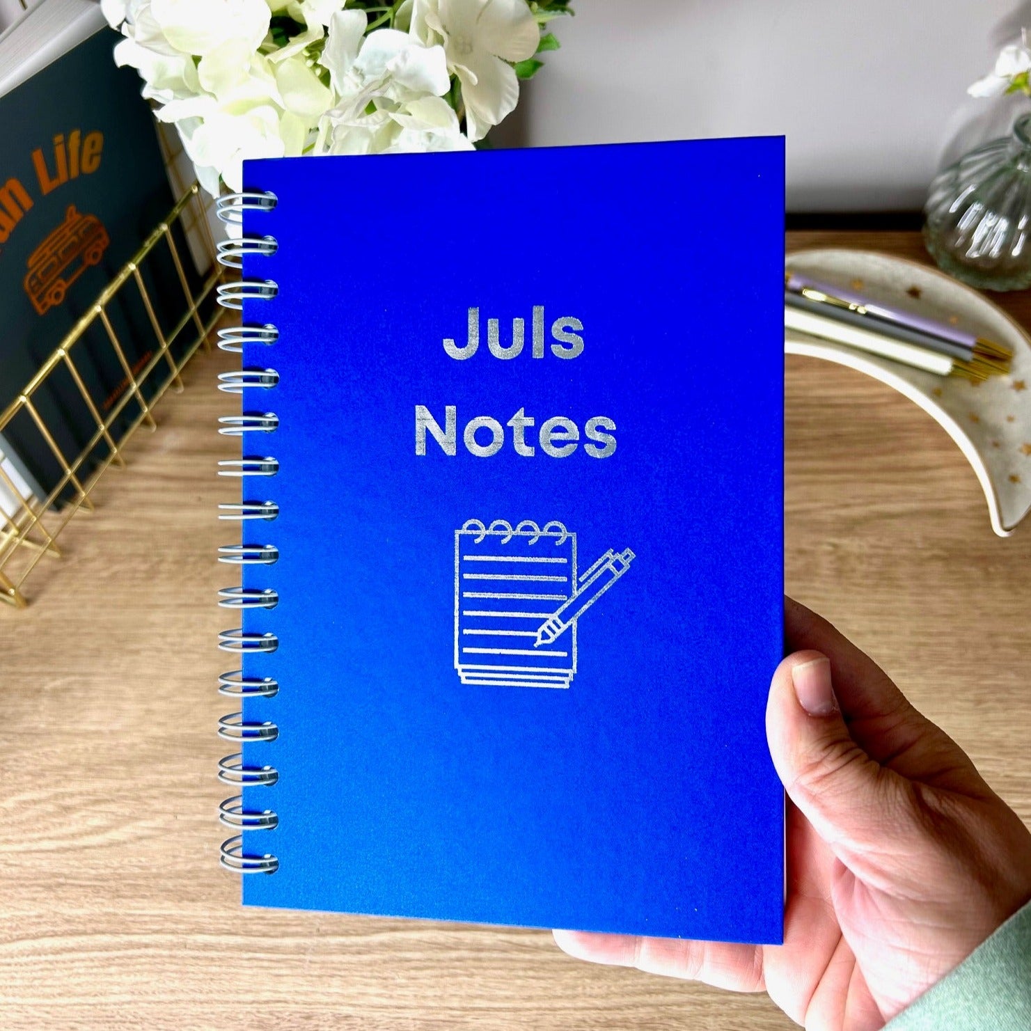 A hardback A5 notebook in Sea Blue that says 'Juls Notes' with an image of a notepad underneath all in silver foil