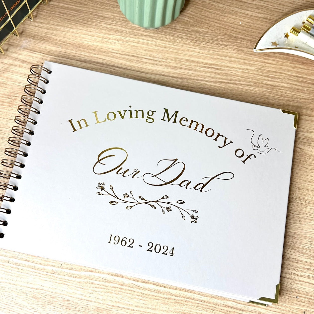 A stone coloured memory book in A4 that says' In Loving Memory of Our Dad 1962-2024' in gold foil and an image of a leaf vine