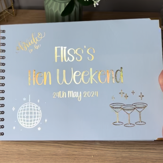 An A4 ice blue memory book with the words 'Fliss's Hen Weekend 24th May 2024' with images of a disco ball and some cocktail glasses all in gold foil