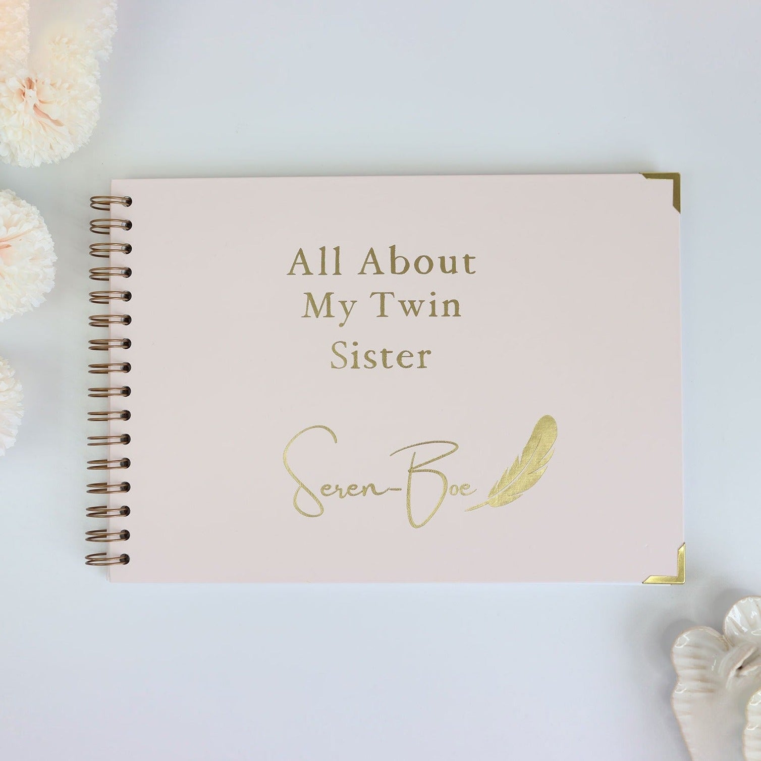 A a4 memory book cover in Pale pink that says All about my twin sister Seren-Boe in gold foil with a gold feather image