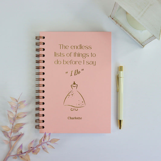 An A5 hardback Powder Pink notebook with the words 'The endless lists of things to do before I say I do' with an image of a wedding dress and the name Charlotte underneath all in gold foil