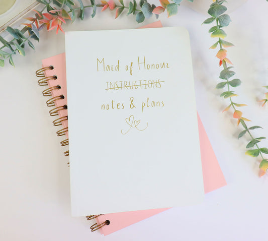 A soft back wite A5 notebook that says 'Maid of Honour (instructions - crossed out) notes and plans' with an image of two hearts in gold foil