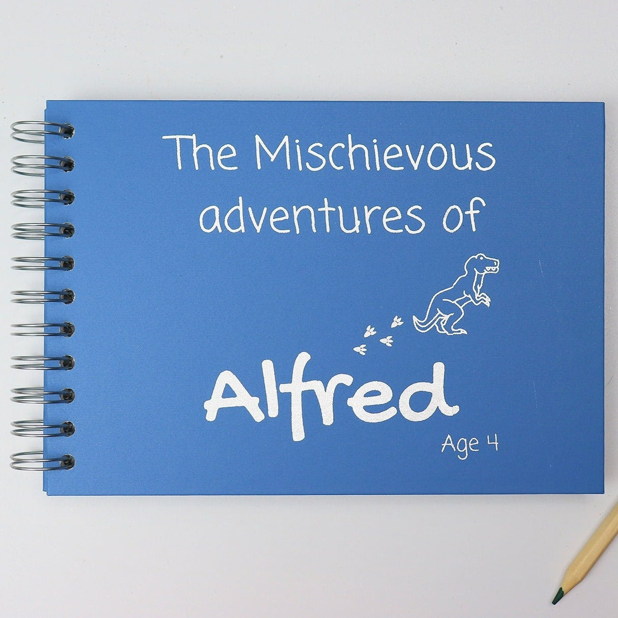 An A5 memory book in Sea Blue with the words 'The Mischievous Adventures of Alfred age 4' with an image of a dinosaur, all in silver foil 
