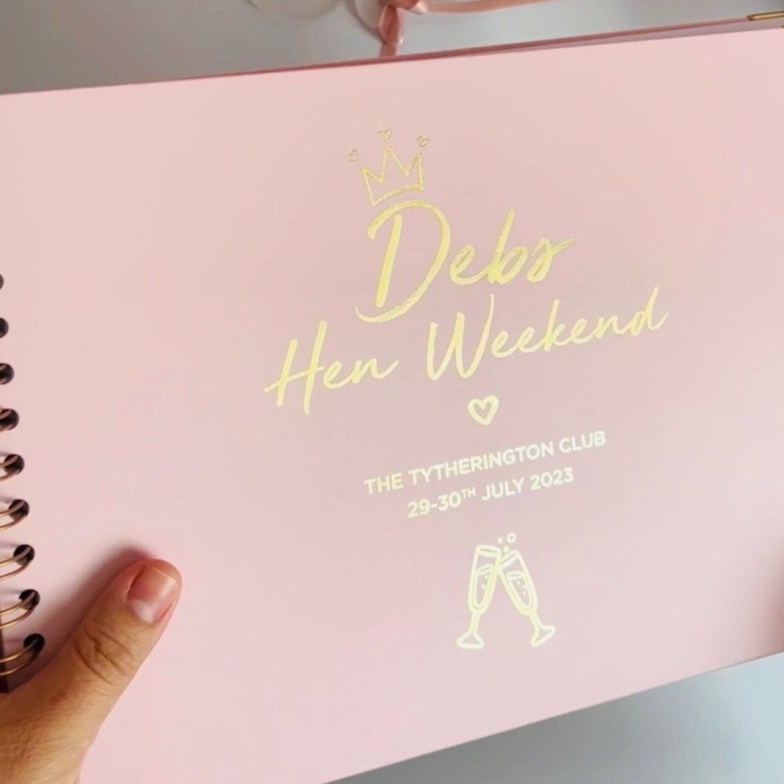 A powder Pink memory book with the words 'Debs Hen Weekend, The tytherington Club' with an image of champagne glasses all in gold foiling
