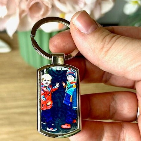 A 7.5cm x 2.5cm metal keyring with a large ring. The surface of the keyring has a pictures of two children smiling taking up the surface of the keyring