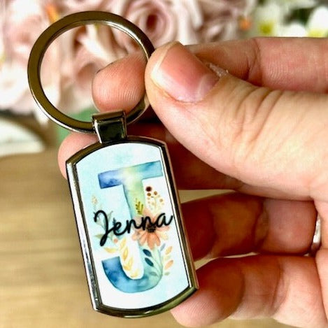A 7.5cm x 2.5cm metal keyring with a large ring. The surface of the keyring has a floral initial of 'J' with the name 'Jenna in handwriting font over the initial taking up the surface of the keyring