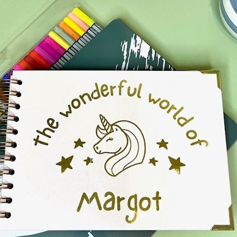 An a5 memory book in pale pink that says 'The wonderful world of Margot' with an image of a unicorn and stars all in gold foil