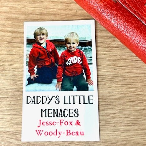 An 8x5 x 5x5 thin plastic card printed in a ploraid style with a picture of 2 small boys and the caption 'Daddy's little Menaces'  and the names of two children underneath