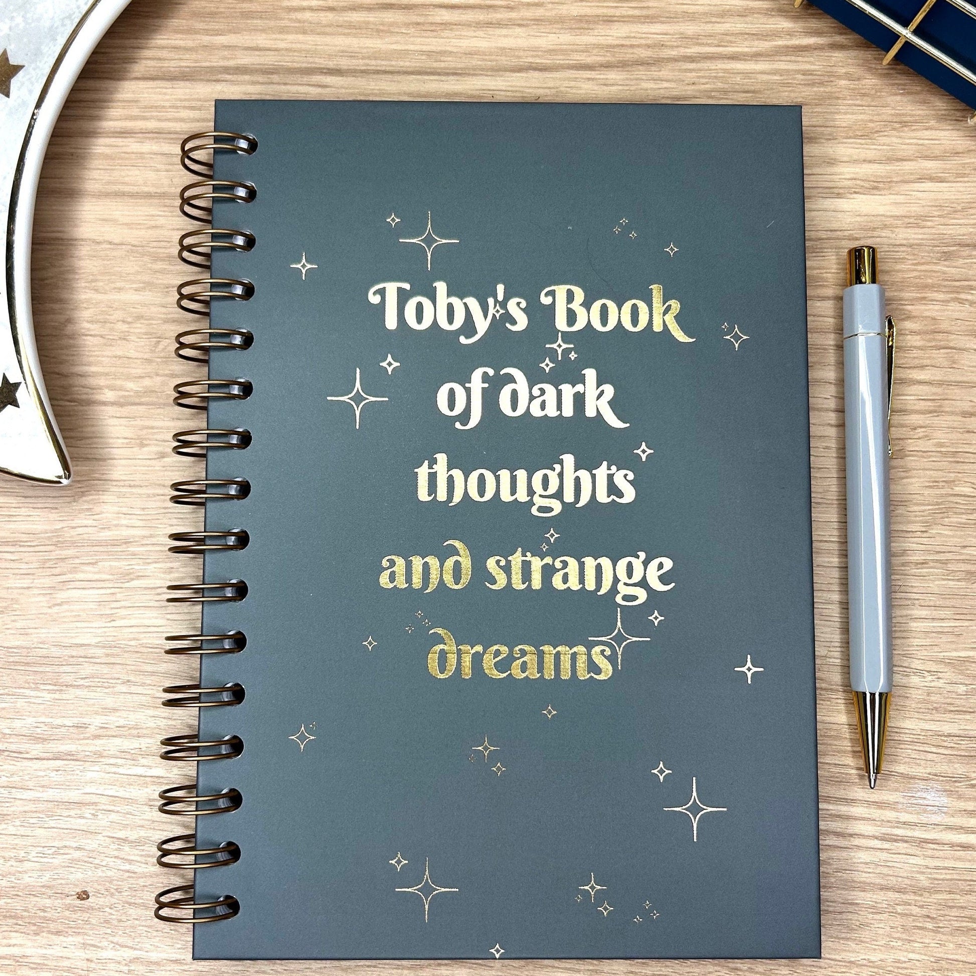 A hardback A5 notebook in Charcoal that says 'Tobys book of dark thoughts and strange dreams' with stars around it all in gold foil