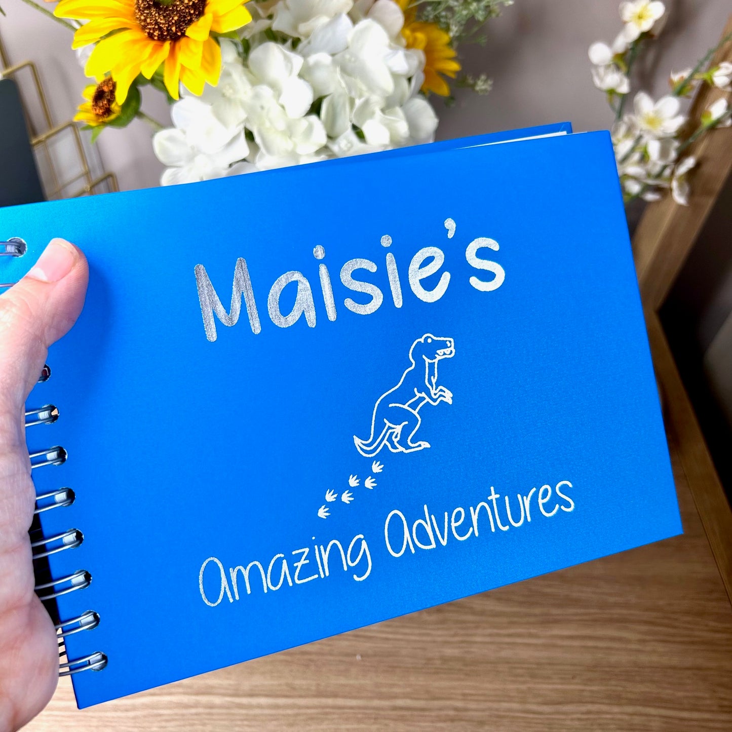 An A5 memory book in sea blue with the words 'Maisie's amazing adventures' with an image of a T rex all in silver foil