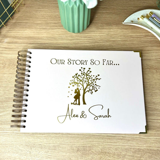 An A4 memory book in Pale pink with the words 'Our story so far...' with an image of a couple kissing below a tree with love hearts for leaves and the names 'Alex and Sarah' underneath, all in gold foil