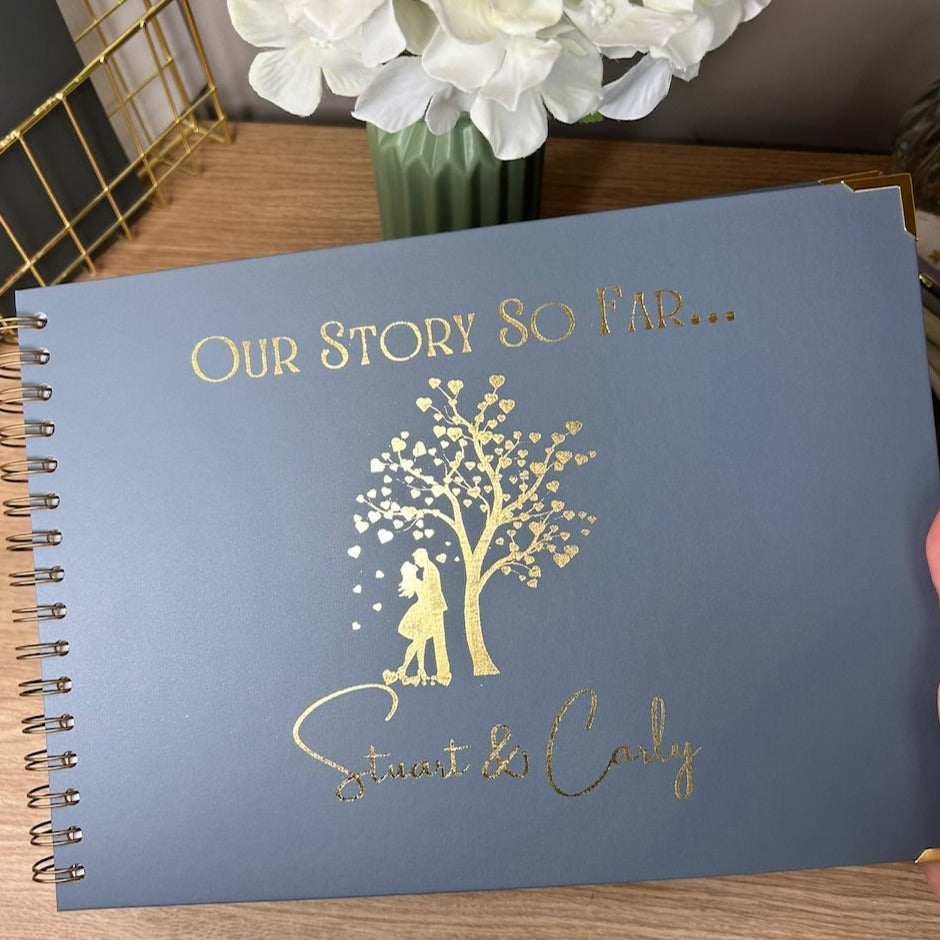 An A4 memory book in Blue/Grey with the words 'Our story so far...' with an image of a couple kissing below a tree with love hearts for leaves and the names 'Stuart and Carly' underneath, all in gold foil