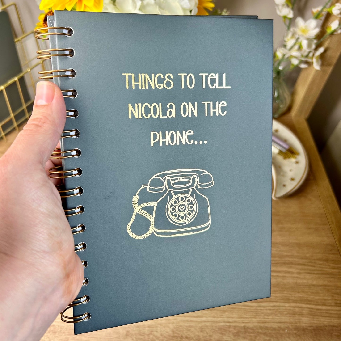 A hardback A5 notebook in Charcoal that says 'Things to tell Nicola on the phone' with and image of a phone underneath, all in gold foil