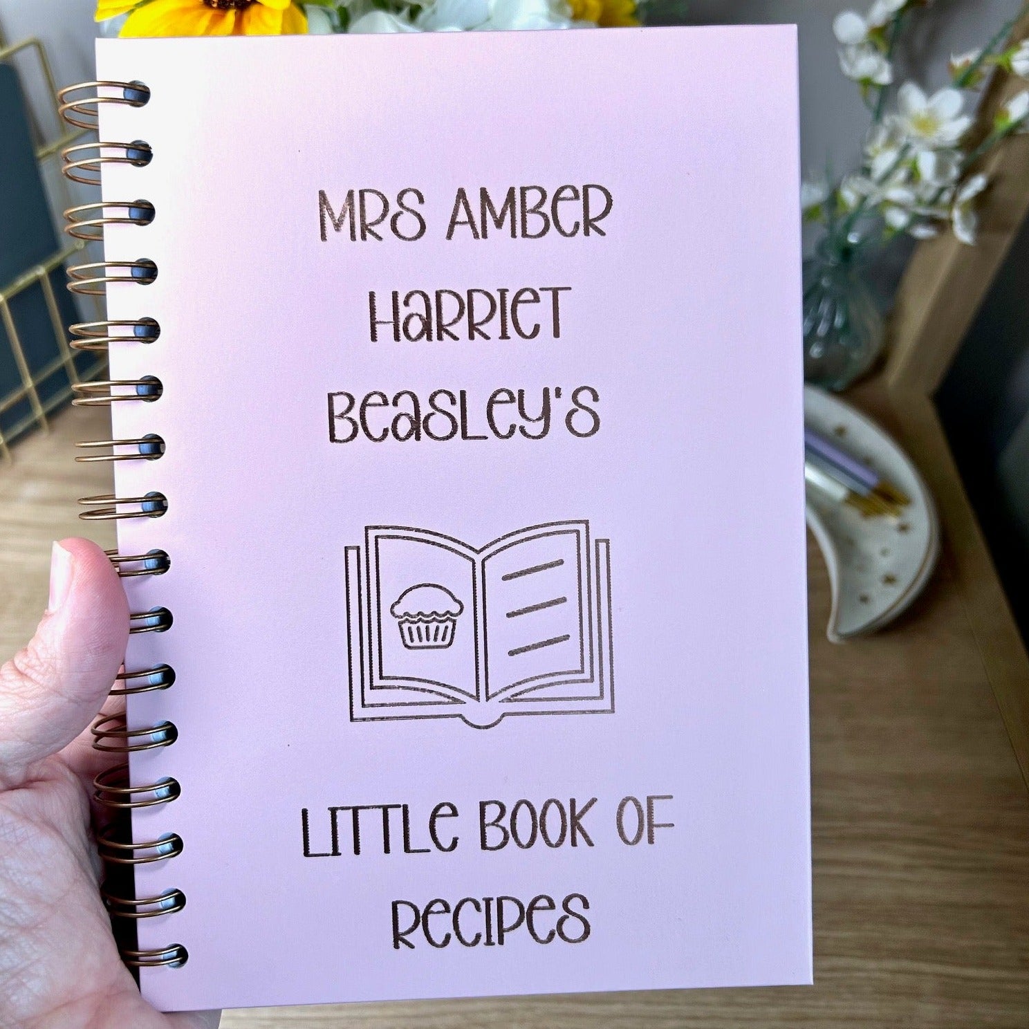 A hardback A5 notebook in powder pink that says 'Mrs Amber Harriet Beasley's little book of recipes' with an image of a recipe book all in gold foil