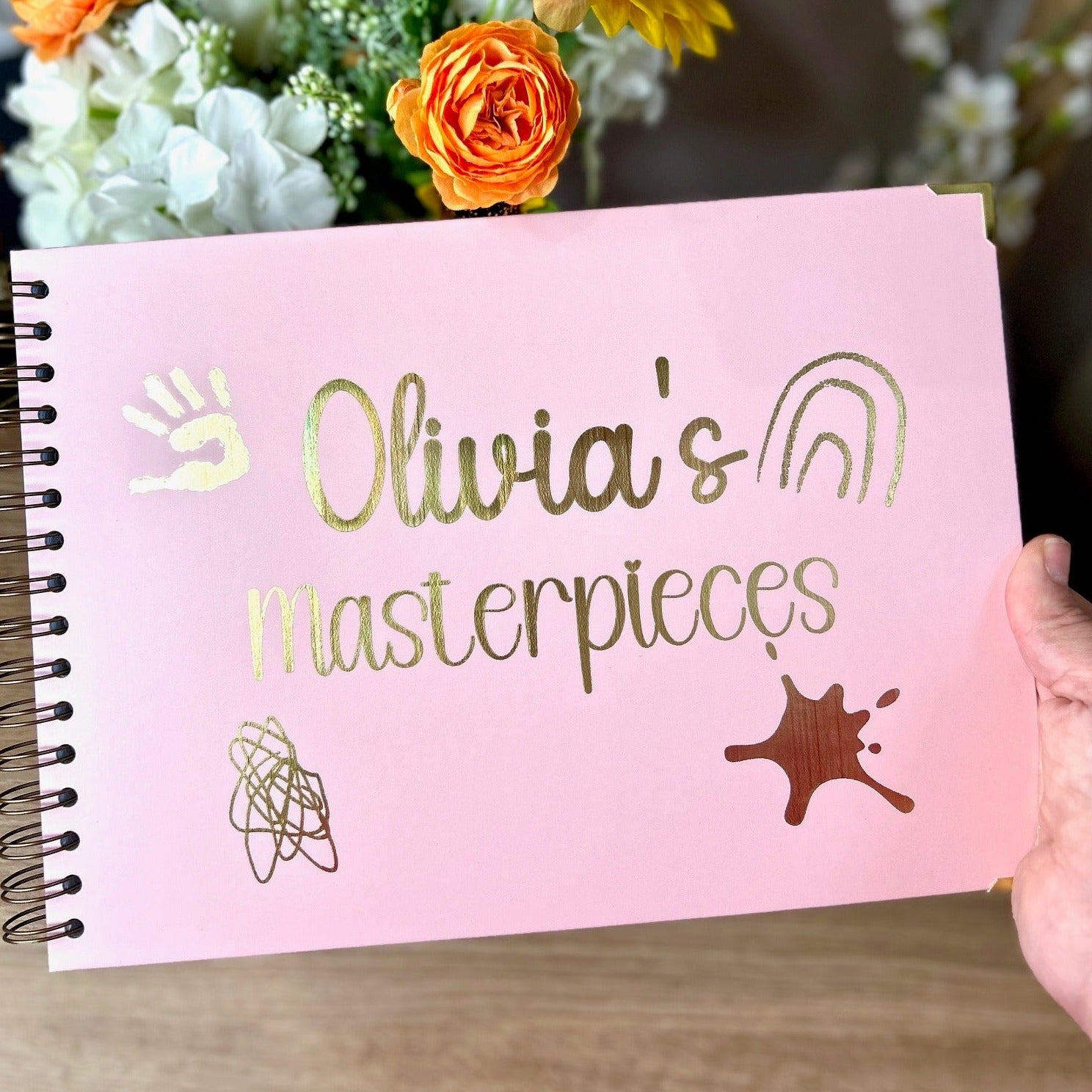 An A4 memory book with the words 'Olivia's Masterpieces' with a paint splodge and some scribbles with a hand print all in gold foil