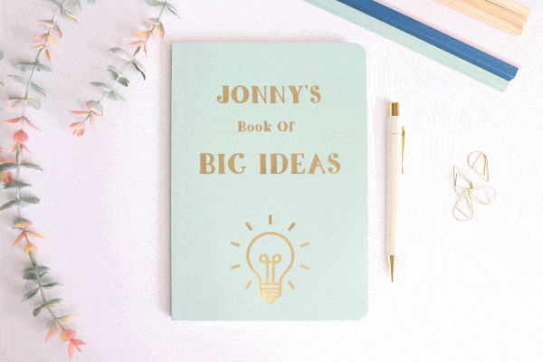 An A5 soft back lined notebook in a Sage colour with the front reading 'Jonny's Book of Big Ideas'. There is a picture of a lightbulb underneath the text which is shining. All writing and pictures are in gold foiling.