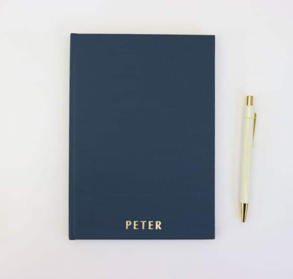 A5 navy blue notebook embossed with the letters PETER lay flat on a white table with a cream and gold hexagonal pen lay next to it
