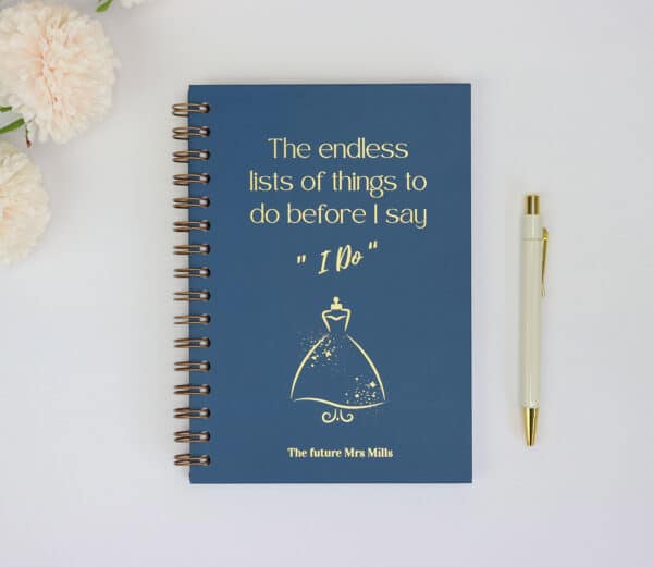 A5 navy blue hardback notebook with the words 'the endless lists of things to do before I say I do, The future Mrs Mills' There is a picture of a wedding dress in the centre of the book. All images and text are in gold foil.