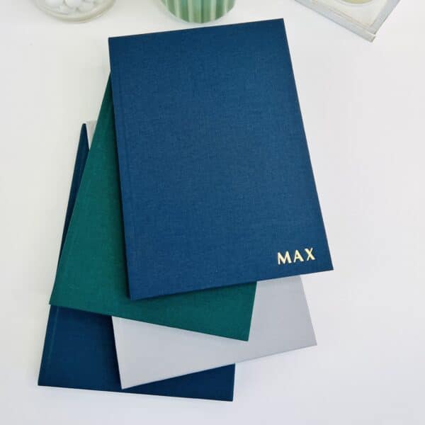 A selection of cotton feel A5 hardback notebooks in the colours emerald green, blue, light grey and purple. The blue book is embossed in gold foil with the word 'max'