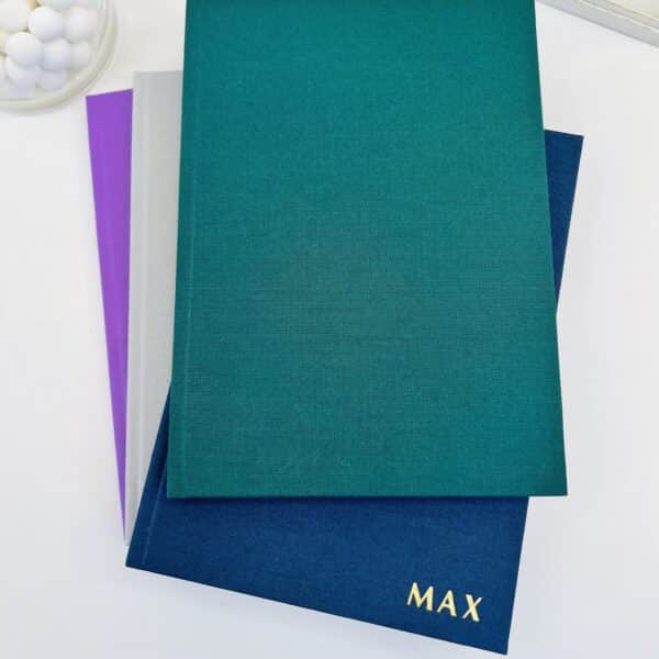 A selection of cotton feel A5 hardback notebooks in the colours emerald green, blue, light grey and purple. The blue book is embossed in gold foil with the word 'max'