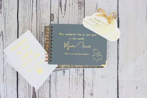 An A5 landscape memory book in blue grey that says How wonderful life is now your in the world Montie Forest in gold foil. A white perspex hanging cloud that is personalised in gold foil with babys name and a card and envelope with baby's name on it.